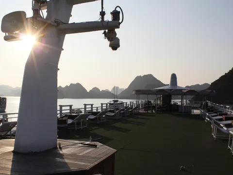 Sun-lit sundeck of a cruise boat with lounge chairs, a shaded bar area and a mast with a camera, looking out over calm water toward limestone karst hills and another boat.
