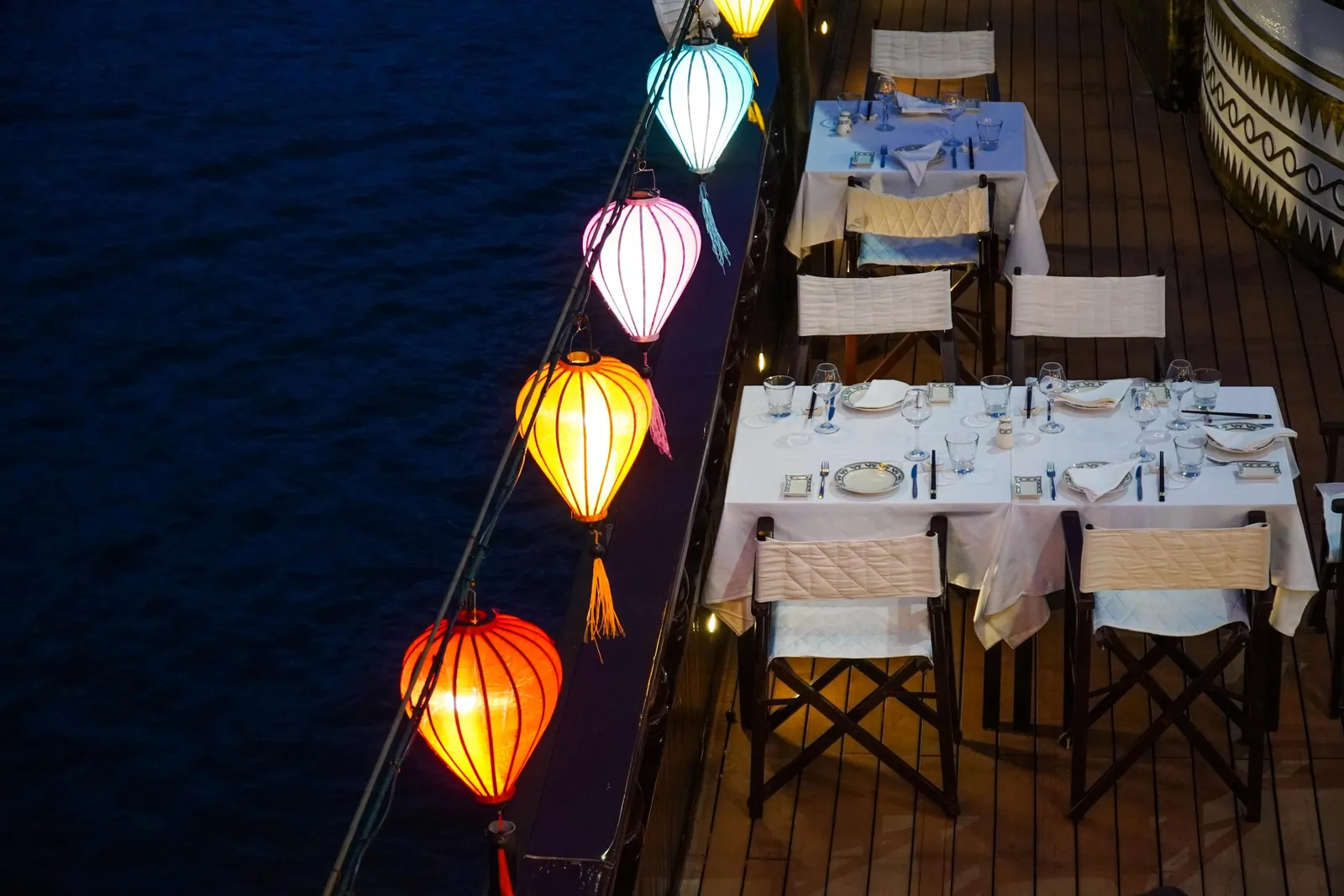 Elevated night view of a wooden boat deck with set white-clothed dining tables and folding chairs beside a railing strung with glowing colorful silk lanterns, next to dark water.