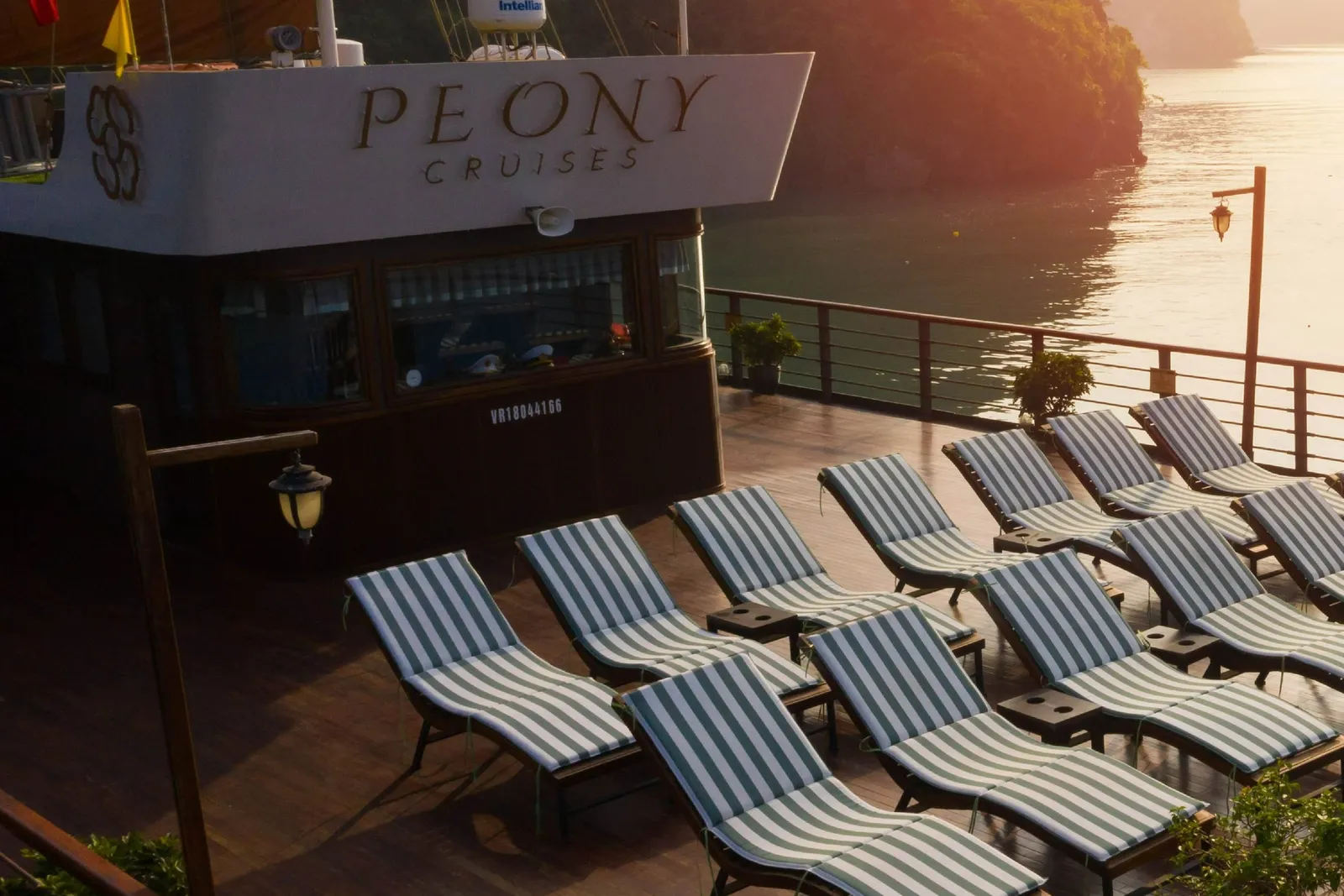 Rows of green-and-white striped sun loungers on a wooden upper deck at sunset, with the wheelhouse bearing 'Peony Cruises' lettering, a Vietnamese flag, and limestone karst islands over calm water behind.