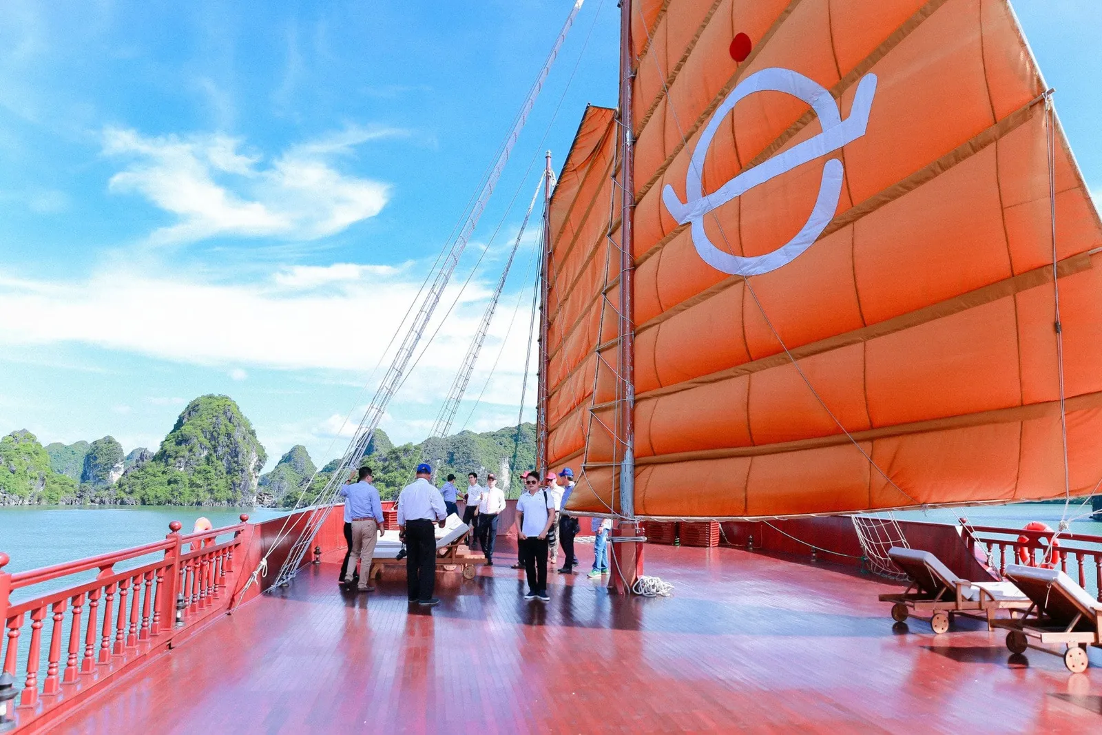 A group of people stands on the red wooden deck of a junk boat with a large orange sail bearing a white logo, with green limestone karst islands and blue sky in the background.