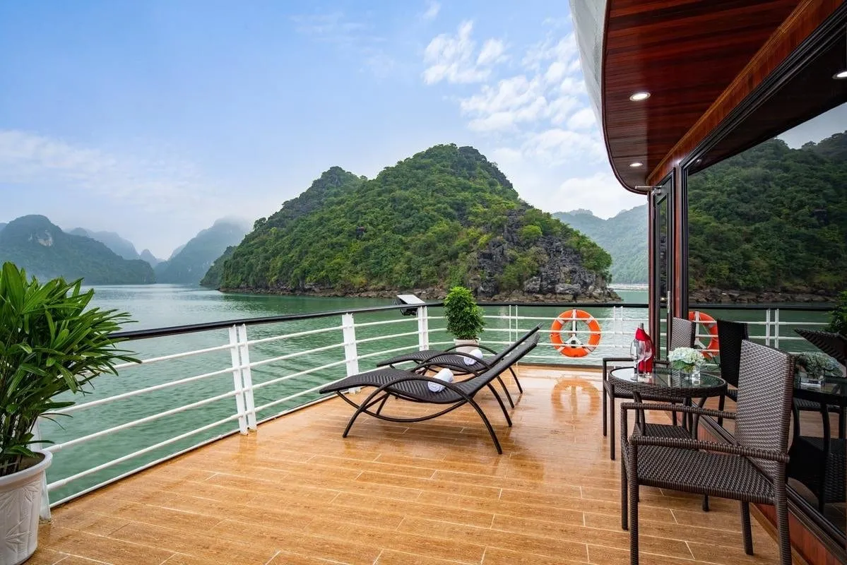 A wooden-floored boat deck with lounge chairs, a small table with flowers and glasses, wicker chairs and life rings, overlooking green water and forested limestone karst islands.