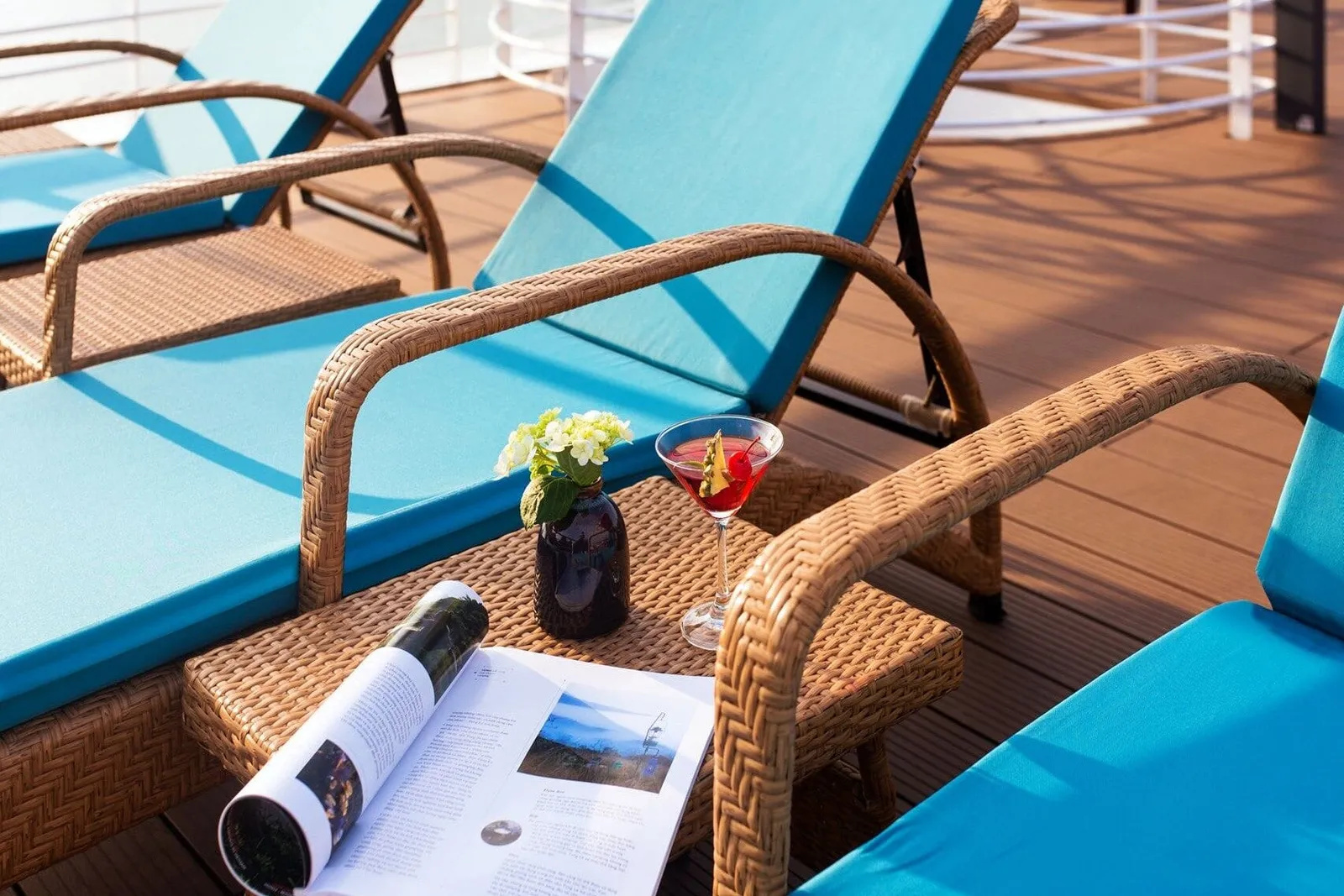 Teal-cushioned wicker sun loungers on a wooden deck with a small wicker table holding a cocktail, a vase of white flowers and an open magazine.