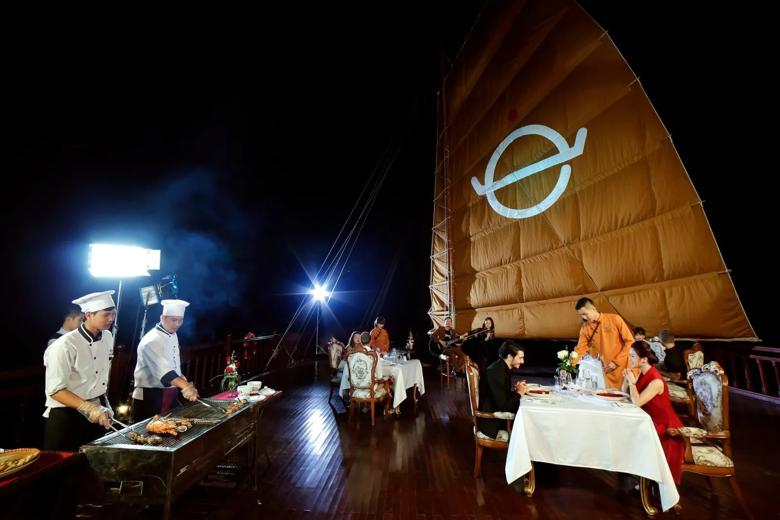 At night on a wooden boat deck beneath a large orange junk sail bearing a white logo, chefs grill seafood at left while a couple dines at a white-clothed table and musicians play in the background.