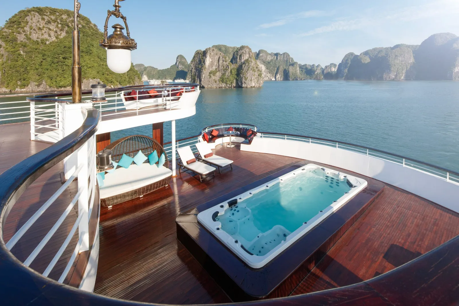 A wooden upper deck of a cruise boat with a built-in jacuzzi, lounge chairs and a wicker daybed, overlooking calm water and limestone karst islands.