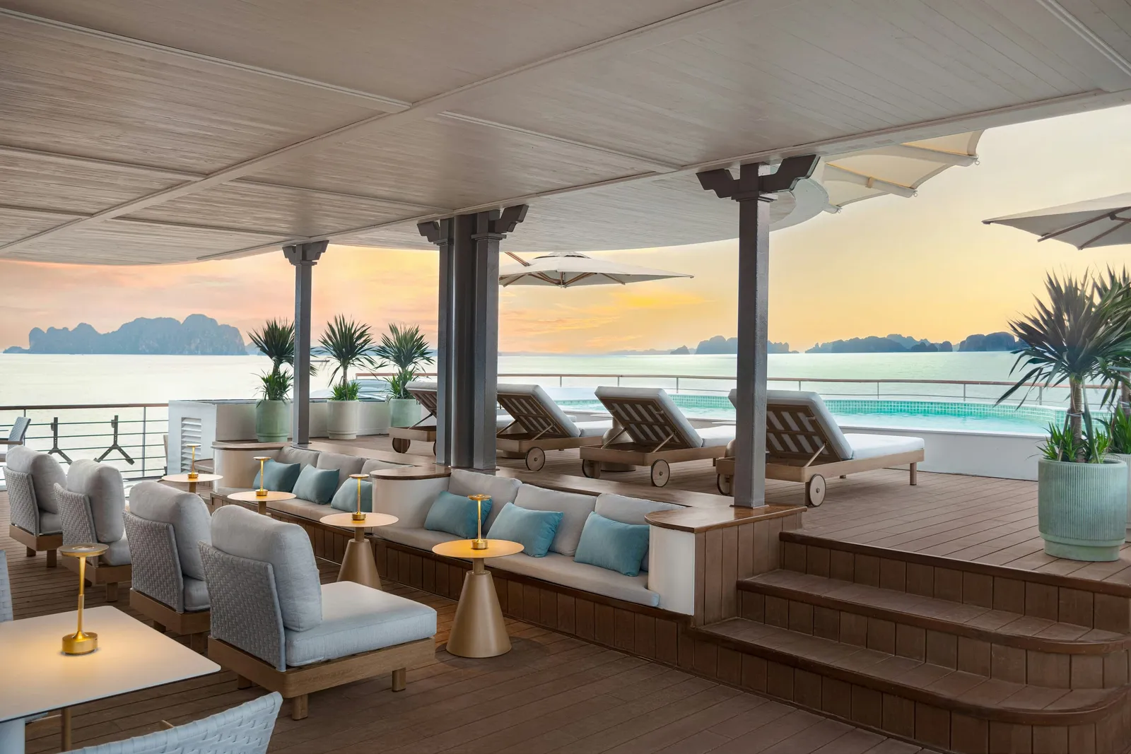 A covered upper deck with lounge seating, sun loungers and a pool, looking out over calm water toward distant limestone karst islands at sunset.