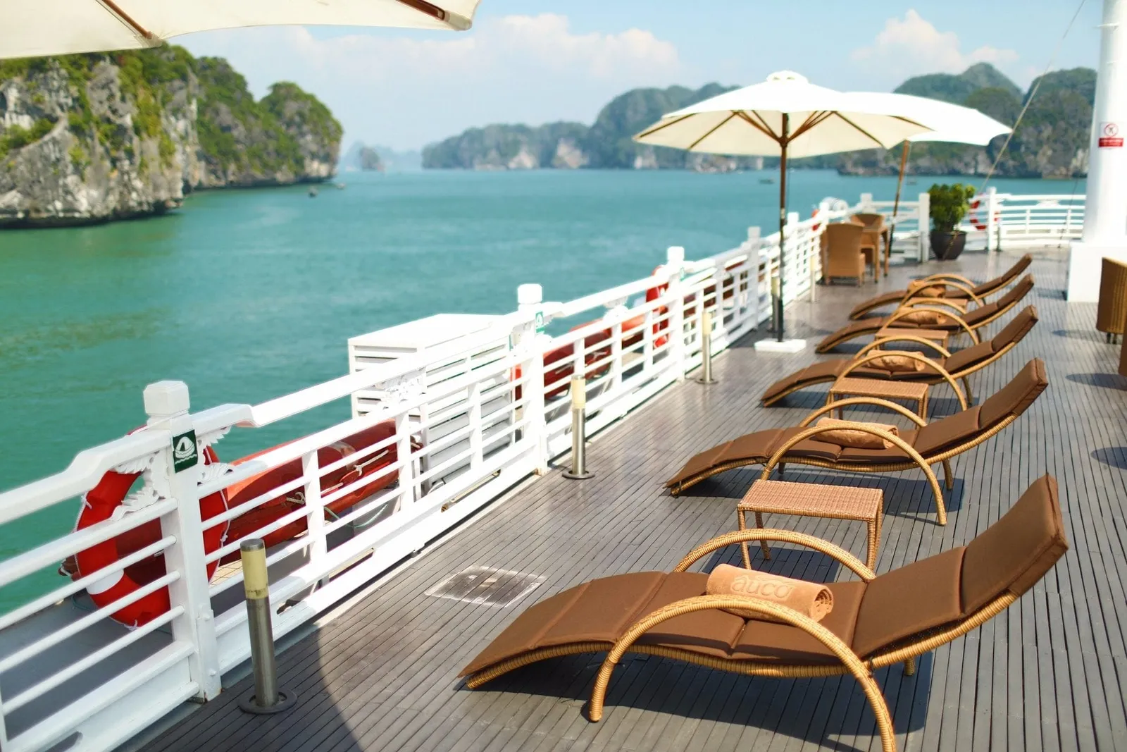 A wooden sun deck with brown wicker loungers, white umbrellas and a white railing with life rings, overlooking turquoise water and green limestone karst islands, with a logo printed on a rolled towel on the nearest lounger.