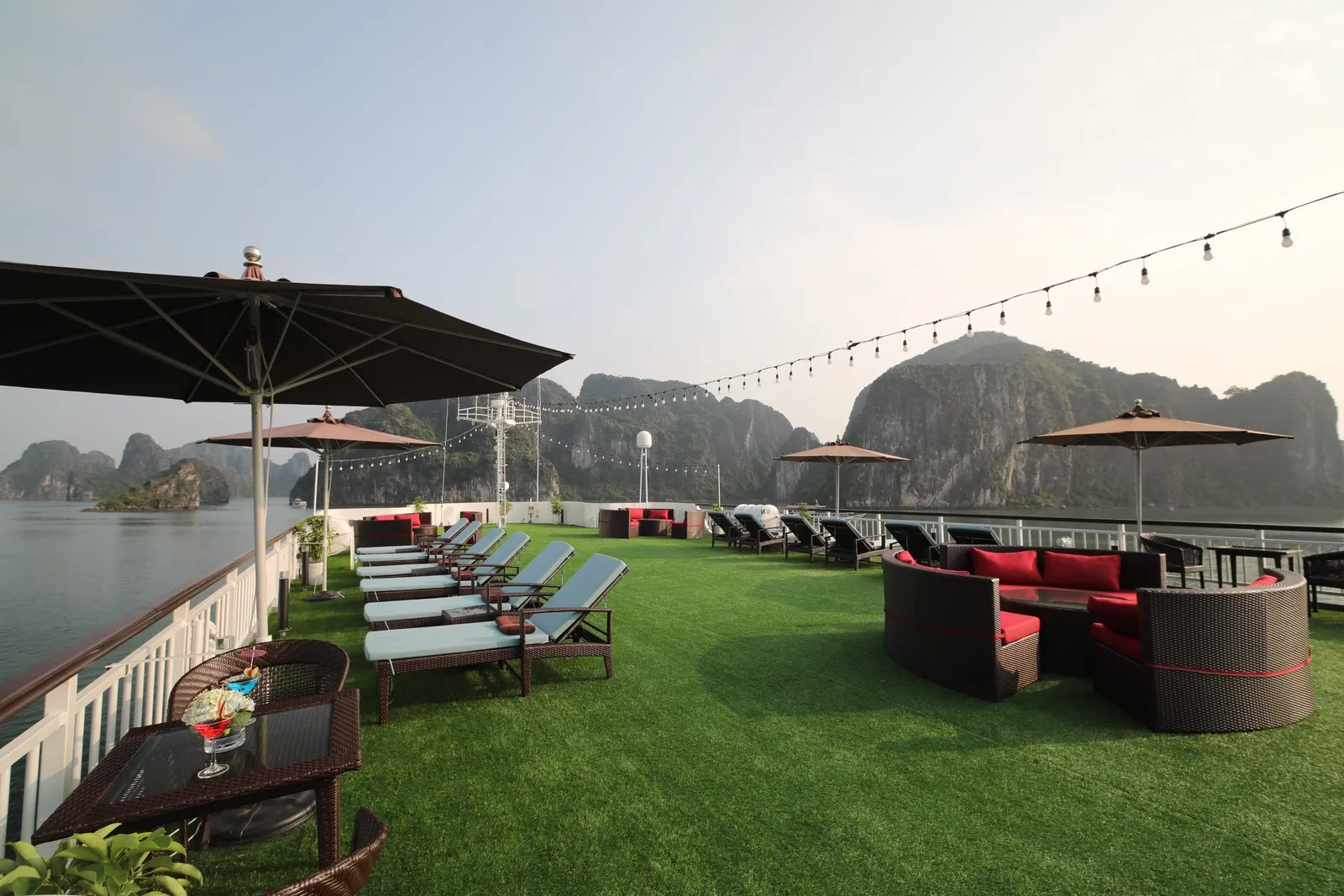 A sun deck covered in artificial grass with lounge chairs, umbrellas, wicker sofas with red cushions and string lights, overlooking calm water and limestone karst islands.