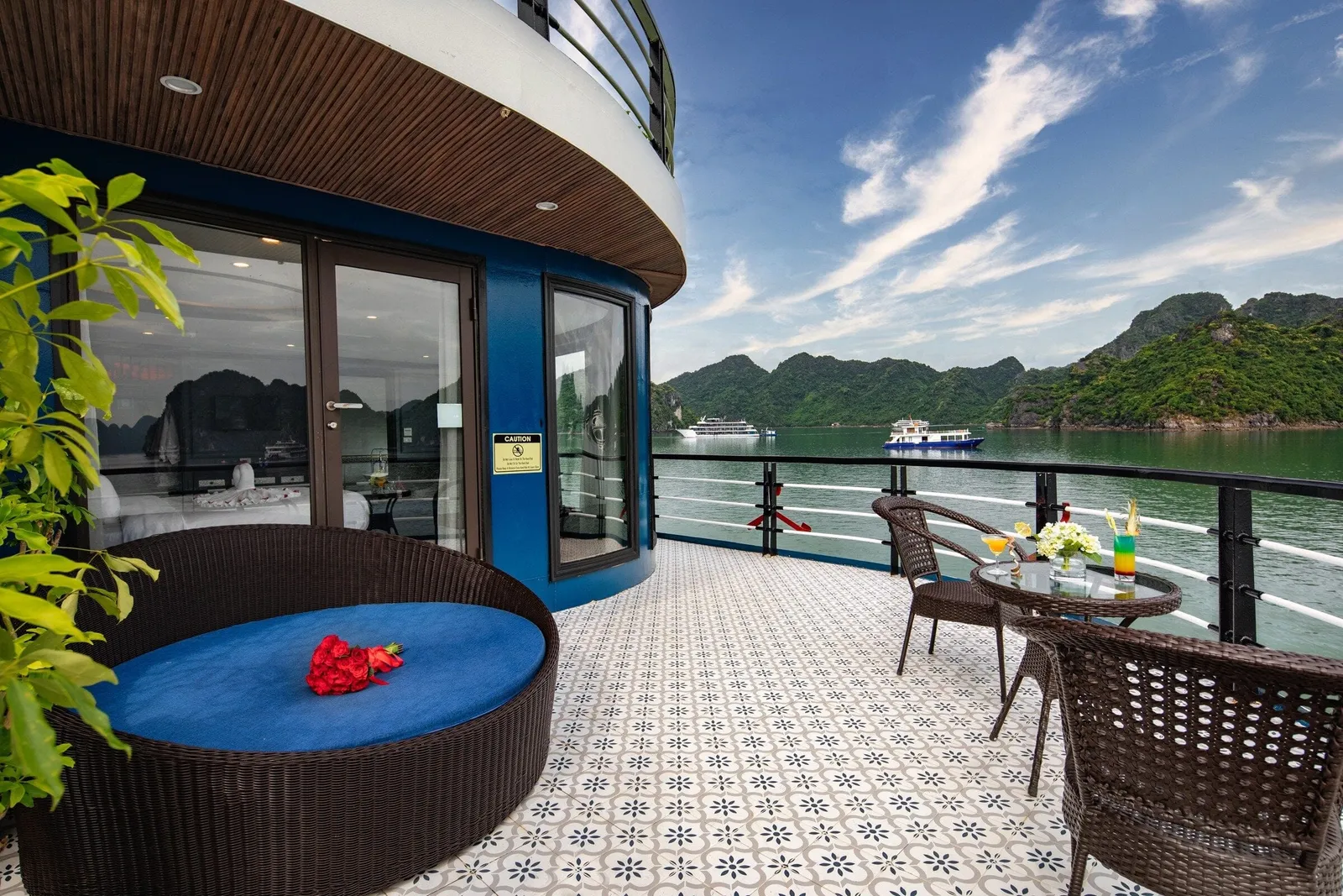 A private balcony deck with patterned tile floor, a round wicker daybed with red roses, a small table with drinks and flowers, and a cabin with glass doors, overlooking green karst islands and other boats on the bay.
