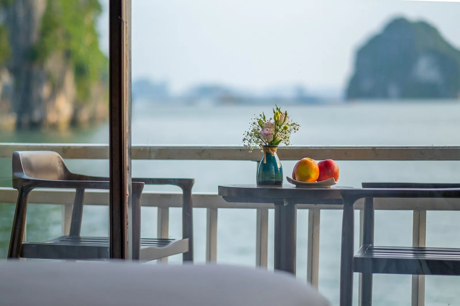 A balcony table with a small flower vase and two peaches on a plate, flanked by chairs, seen through a glass door toward calm water and limestone karst islands.