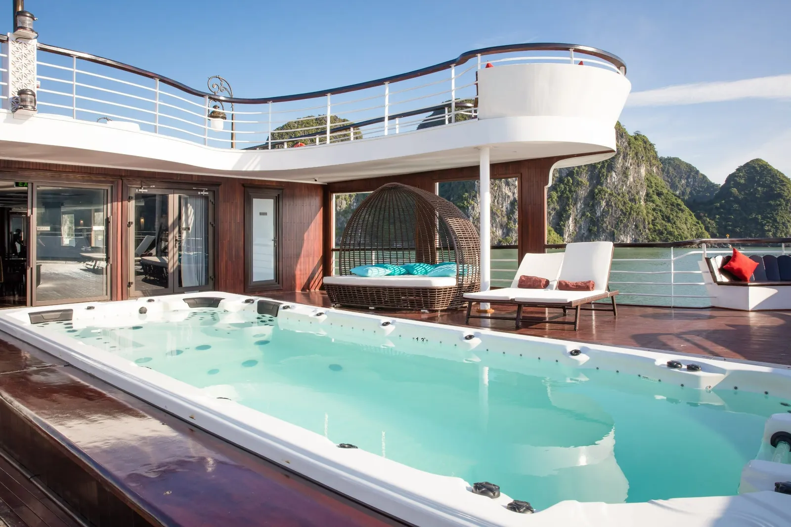 A sun deck on a cruise boat with a jetted plunge pool, a wicker daybed, white loungers and a curved upper deck railing, with green limestone karst cliffs and calm water behind.