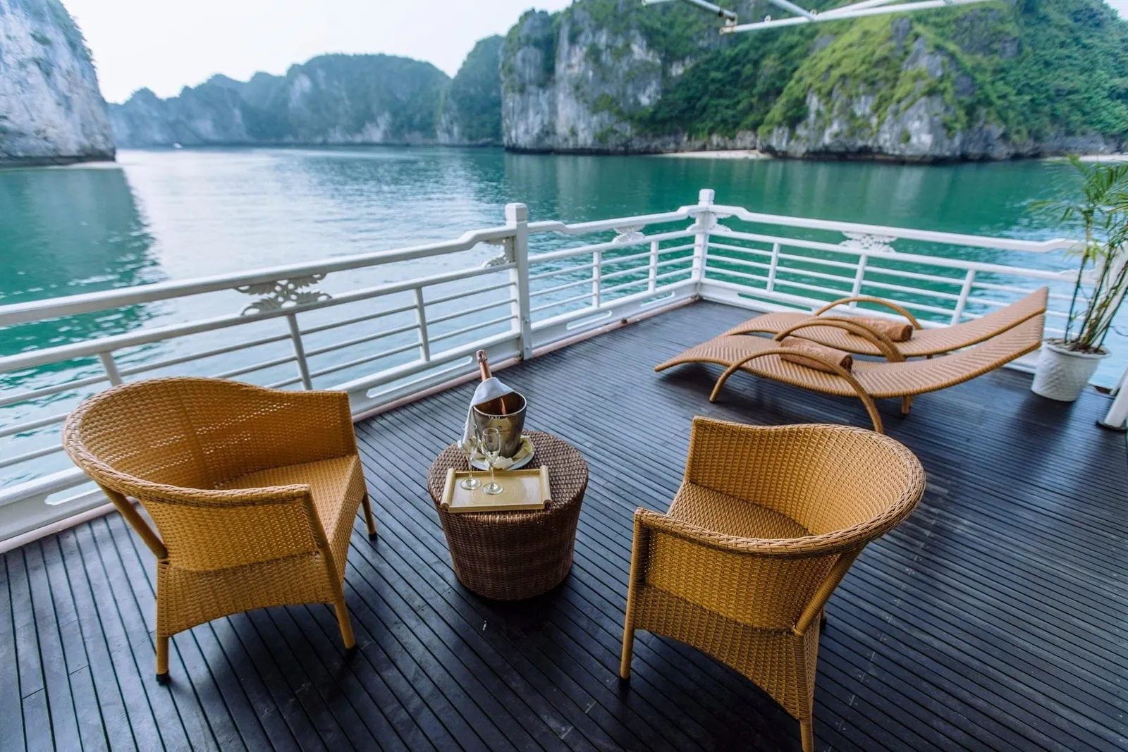A dark wood boat deck with wicker chairs, a small table holding a champagne bucket and glasses, two loungers and a white railing, overlooking green water and tree-covered limestone karst cliffs.