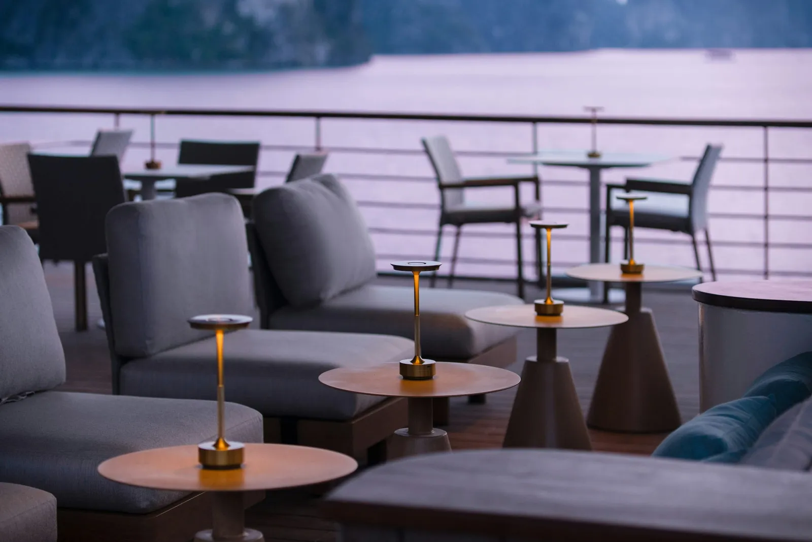 A dusk view of an outdoor boat deck lounge with grey cushioned sofas, small round tables with lit brass lamps, chairs behind a railing, and calm water with hazy karst-like hills beyond.