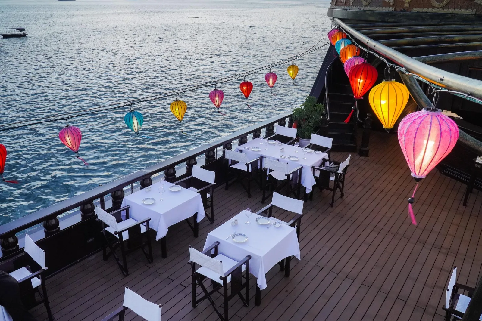 A wooden boat deck set with white-clothed dining tables and folding chairs, with colorful silk lanterns hanging along the railing above open water and a small boat in the distance.