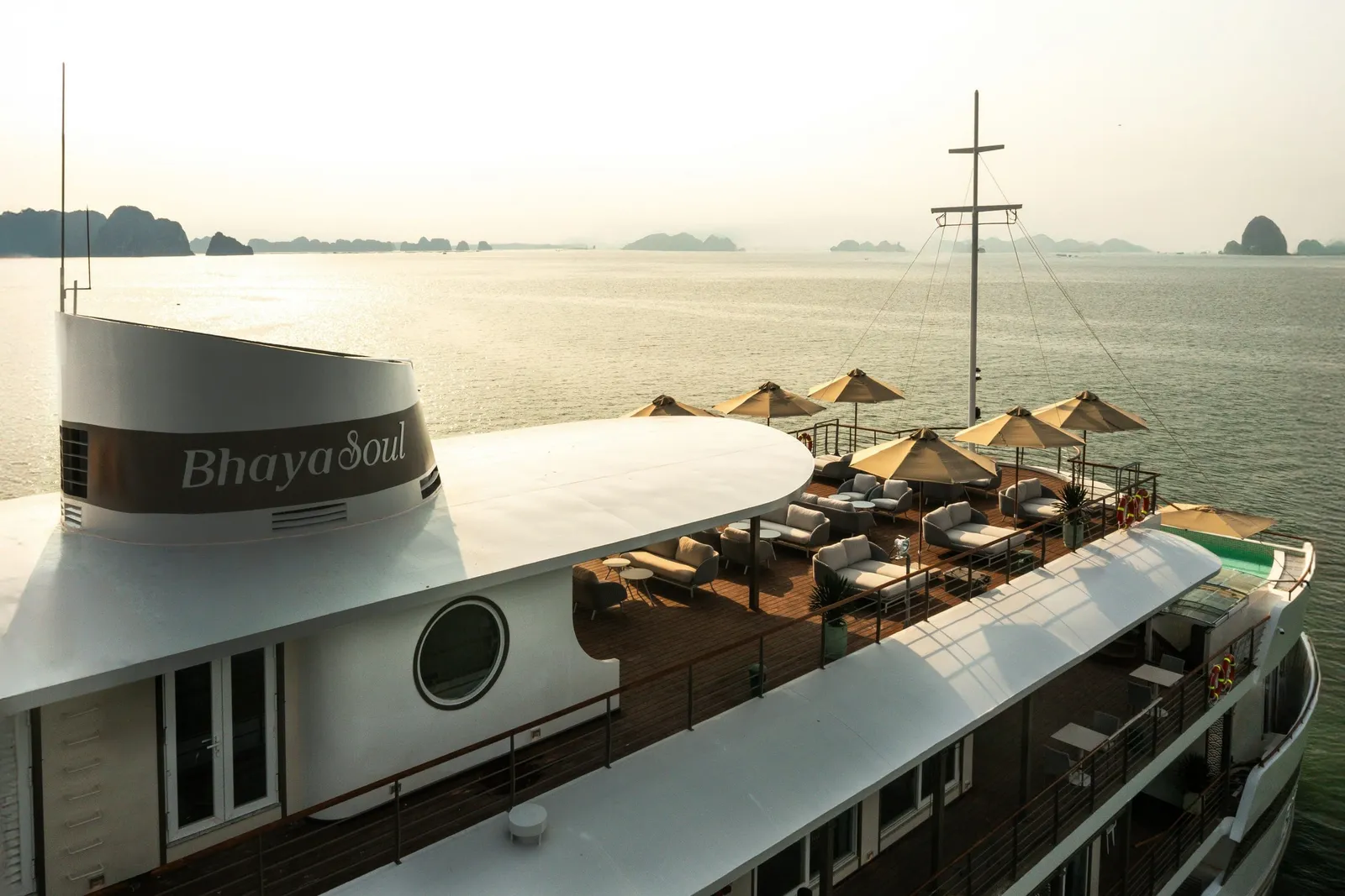 Elevated view of a cruise ship's upper sun deck with lounge seating and tan umbrellas, with 'Bhaya Soul' painted on the funnel and limestone karst islands in hazy water behind.