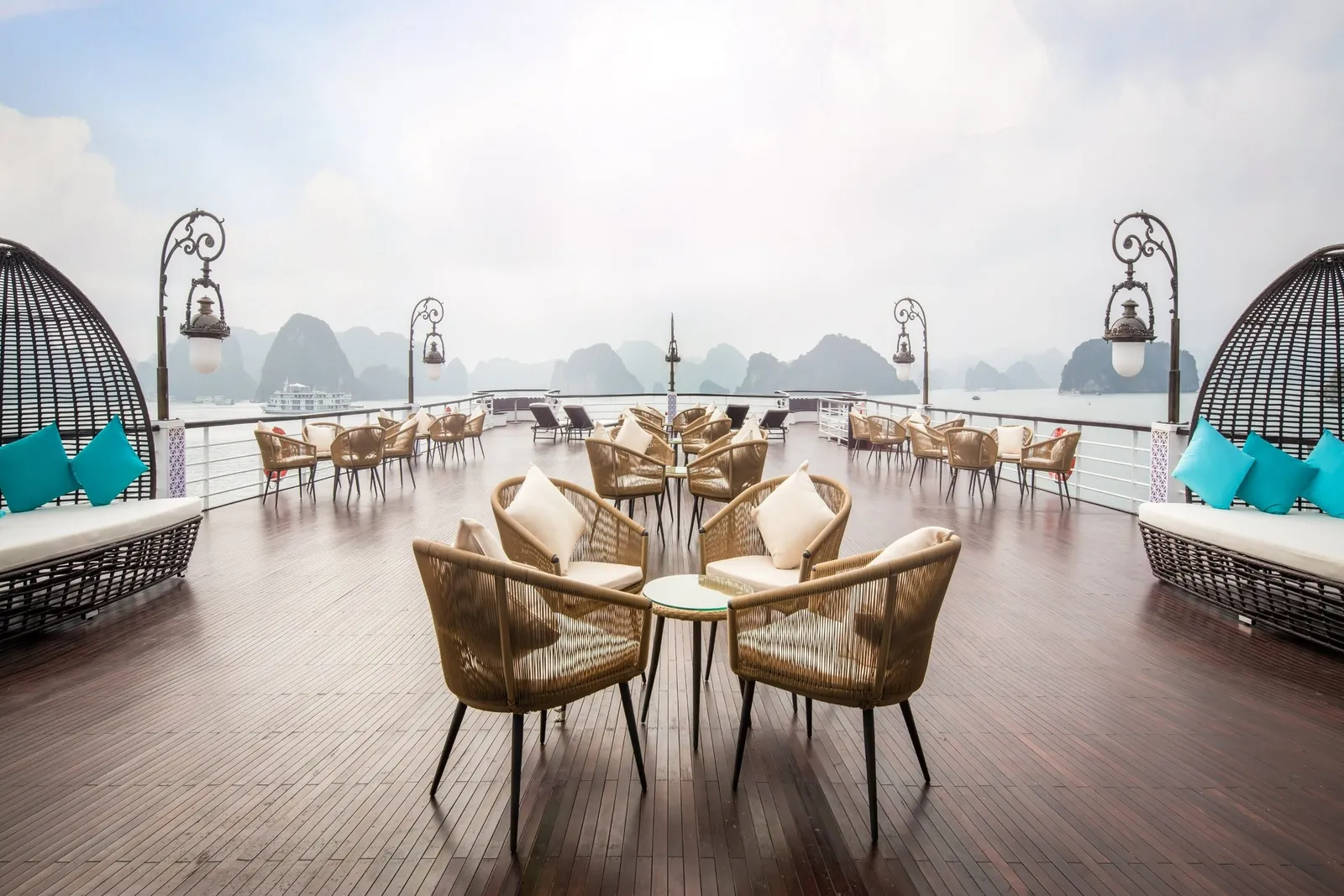 A wooden upper deck with wicker chairs, small round tables, ornate lamp posts and two egg-shaped daybeds with teal cushions, looking out over misty limestone karsts and a distant white boat.