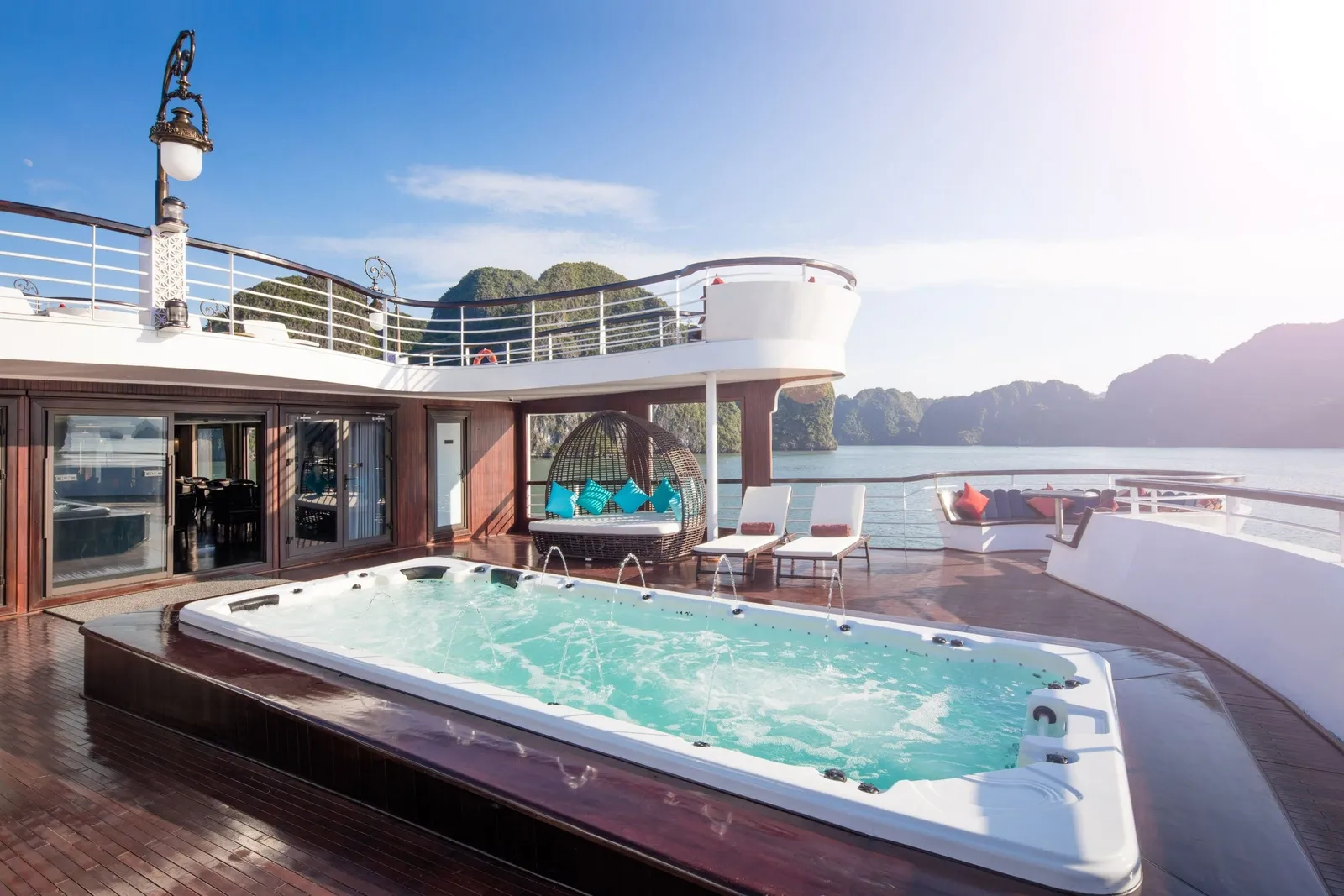 A sun deck of a cruise boat with a built-in jacuzzi pool, a wicker daybed with teal cushions, lounge chairs and seating, with green limestone karst islands and calm water in the background.