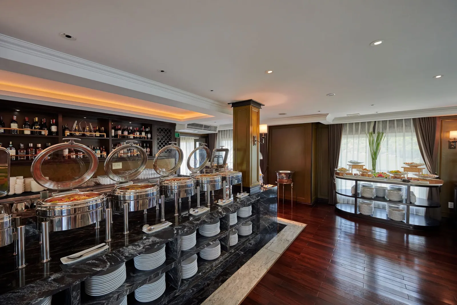 An indoor dining room with a black marble buffet counter holding chafing dishes and stacked plates, a bar shelf of bottles on the left, a second food display on the right, and dark wood floors with curtained windows.