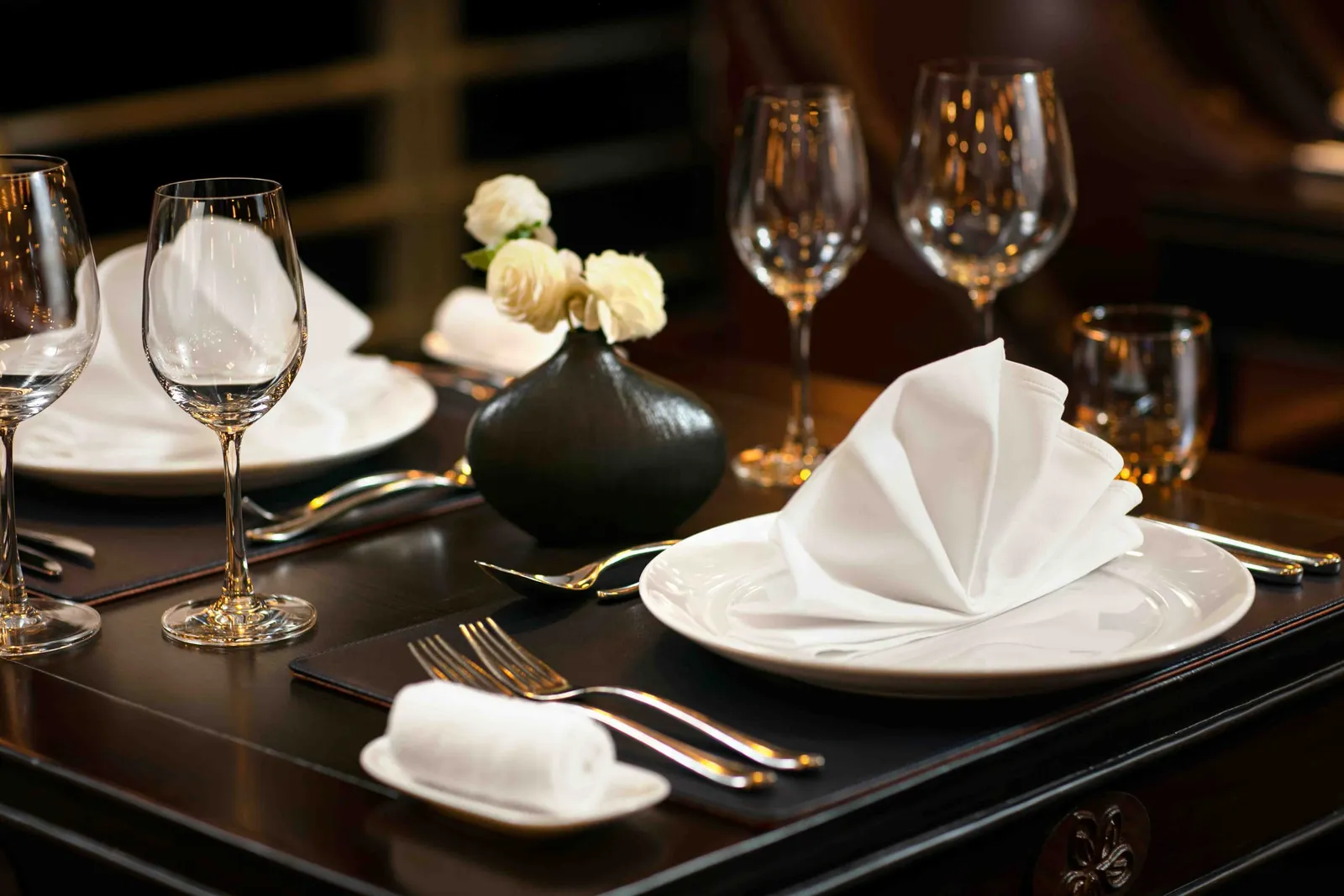A dark wooden dining table set with white plates, folded napkins, wine glasses, cutlery, and a black vase of white roses in a dimly lit room.