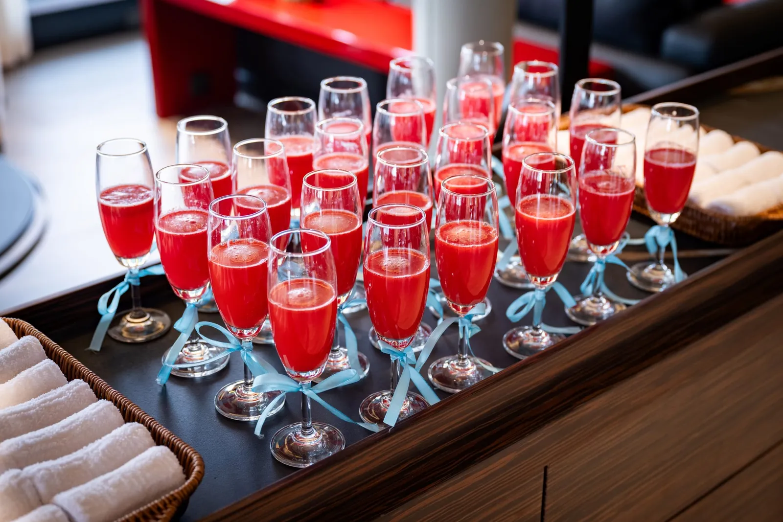 Rows of champagne flutes filled with red welcome drinks and tied with light blue ribbons sit on a dark wooden tray, with wicker baskets of rolled white towels at either side and a red table and black sofa blurred in the background.