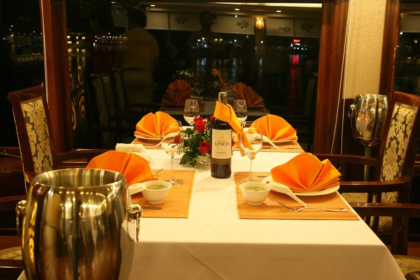 A set dining table in a restaurant with orange folded napkins, a Lynch Bordeaux wine bottle, red roses, soup bowls, wine glasses and steel ice buckets, with windows showing dark night lights behind.