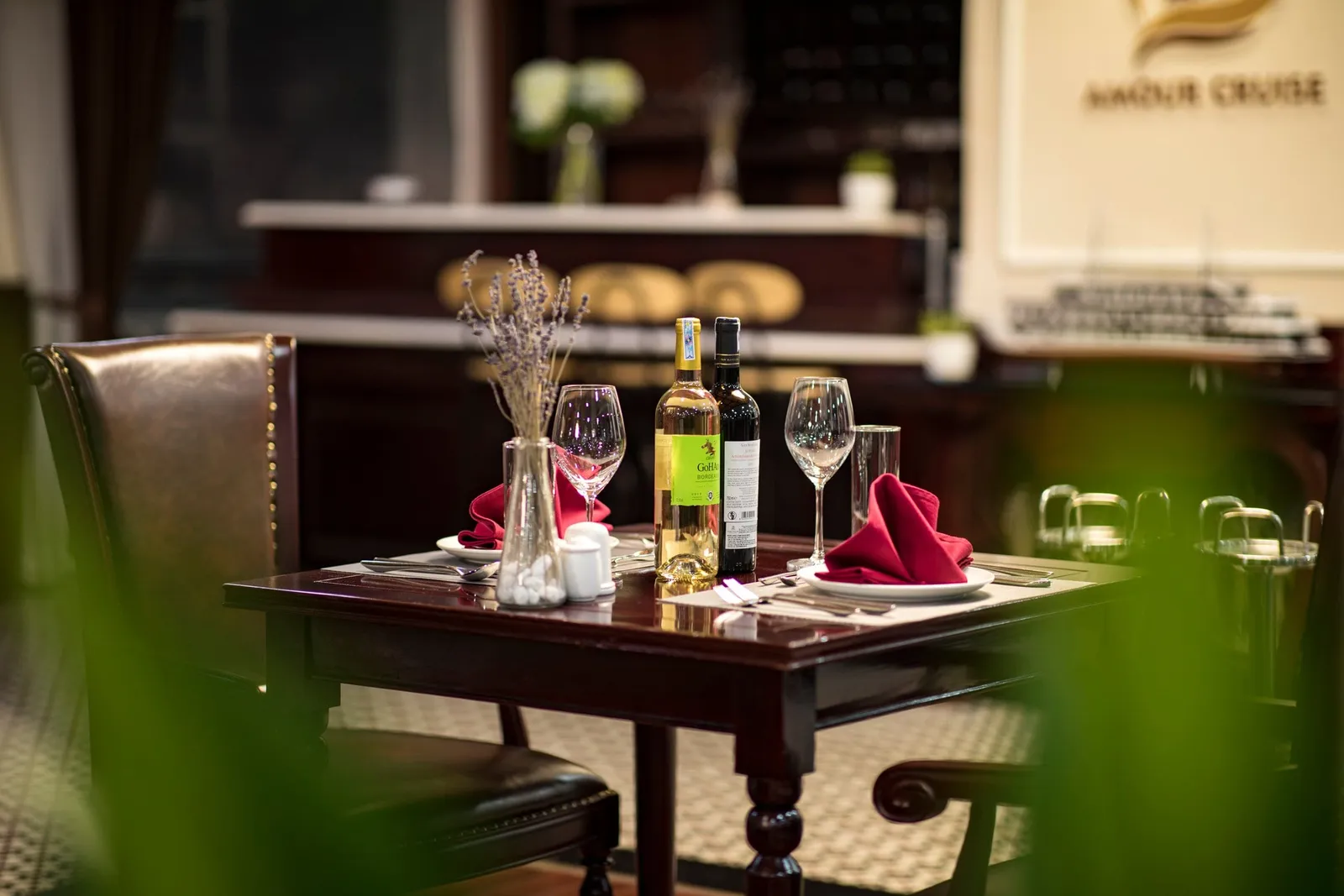 A set dining table with red napkins, wine bottles, glasses and a lavender vase in an indoor restaurant, with a wall sign reading 'Amour Cruise' in the background.