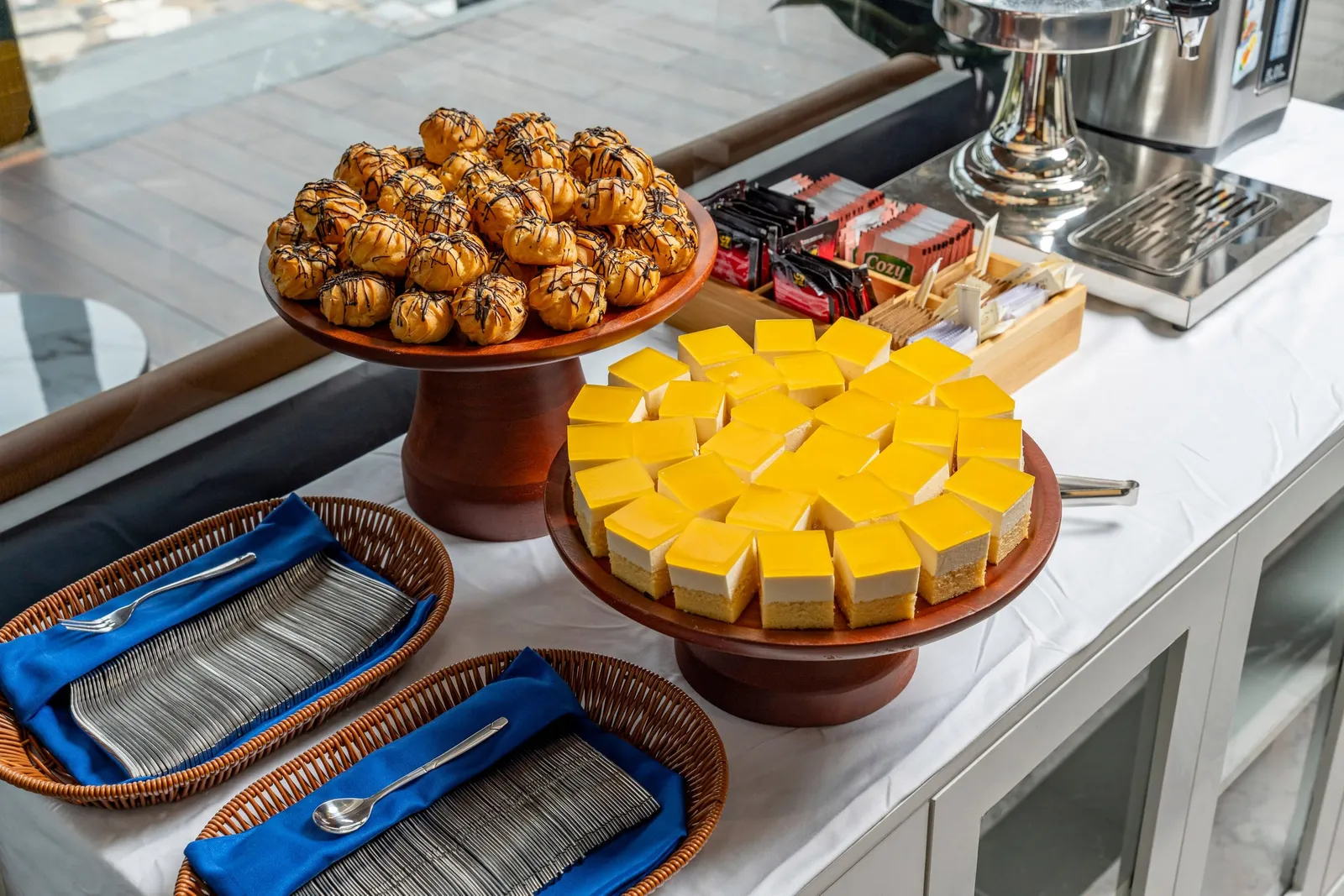 A buffet counter with a white tablecloth holds a stand of chocolate-drizzled cream puffs, a stand of yellow mango cheesecake squares, baskets of forks and spoons on blue napkins, tea packets branded 'Cozy', and a hot water dispenser, with a wooden deck seen through glass behind.