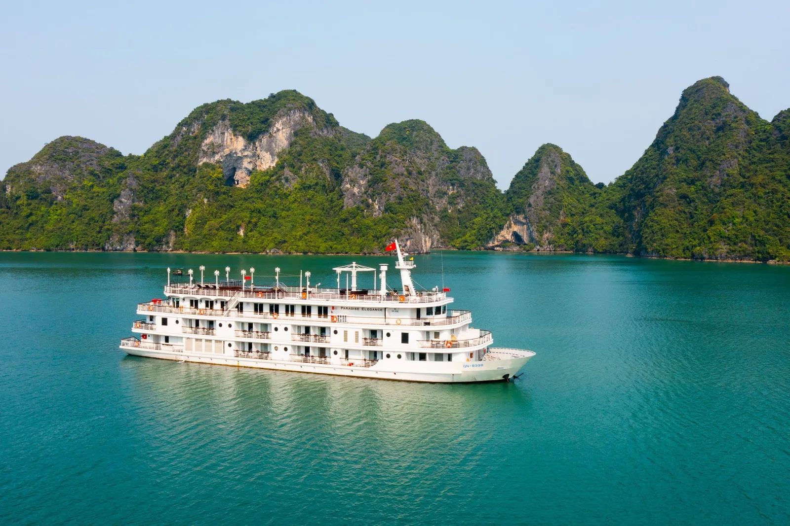 A multi-deck white cruise ship with small name lettering on its side sits on turquoise water in front of green limestone karst islands, photographed from an elevated vantage point off the vessel.