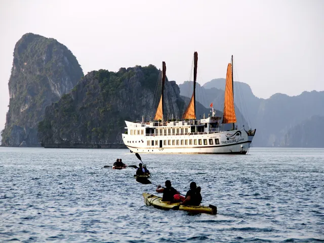 A white three-masted cruise boat with orange sails and 'Treasure Junk' lettering on its hull sits in front of limestone karst cliffs while kayakers paddle in the foreground.