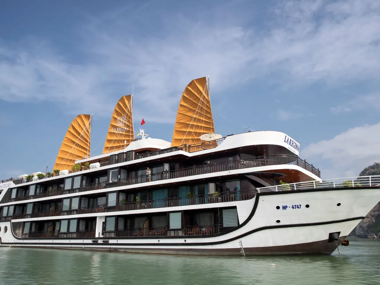 A multi-deck white cruise ship with three orange junk-style sails and 'La Regina' lettering on its hull and sails floats on green water beside a limestone karst under a blue sky with clouds.