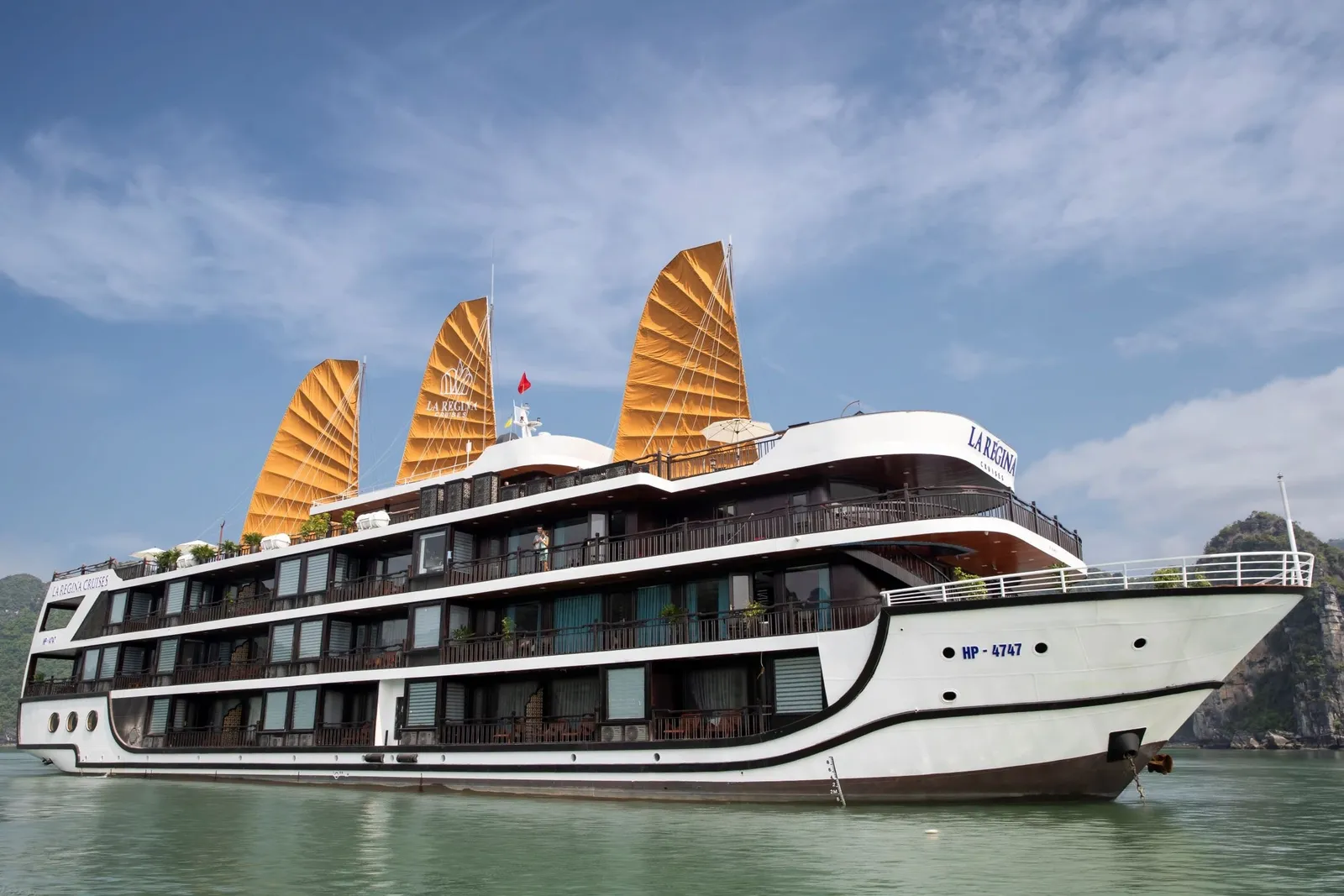A multi-deck white cruise ship with three orange junk-style sails and 'La Regina' lettering on its hull and sails floats on green water beside a limestone karst under a blue sky with clouds.