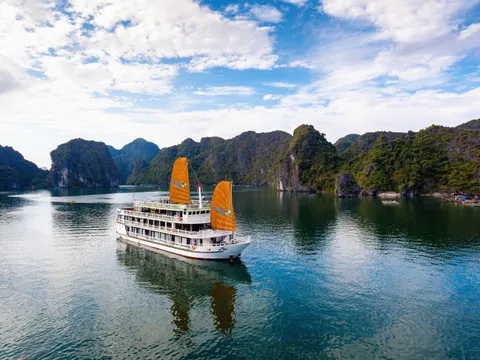 A white multi-deck cruise boat with two orange sails and logos on the sails and hull floats on calm green water in front of forested limestone karsts under a partly cloudy blue sky, photographed from an elevated, drone-like angle.