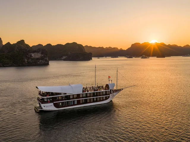 An aerial view of a white and dark-red multi-deck cruise boat with a sail-like canopy and a company logo on its hull, sitting on calm water at sunset among limestone karst islands with other boats in the distance.