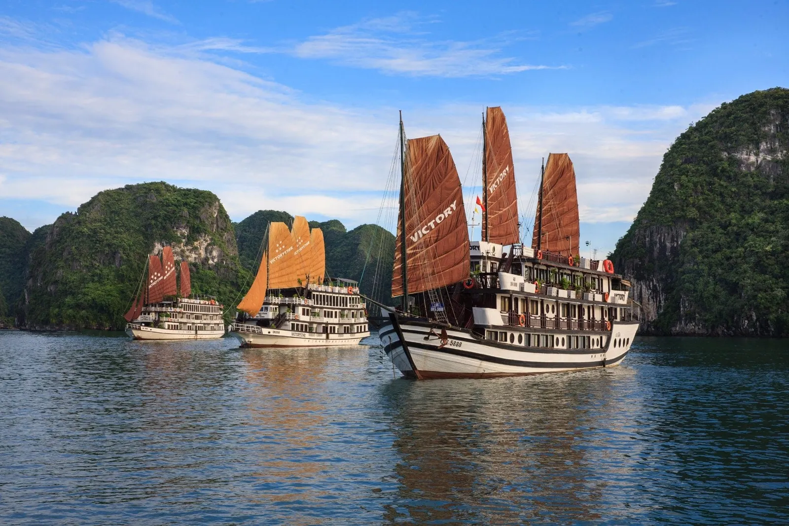 Three multi-deck junk-style cruise boats with brown/orange sails and 'VICTORY' printed on them sail among green limestone karst islands on calm water.