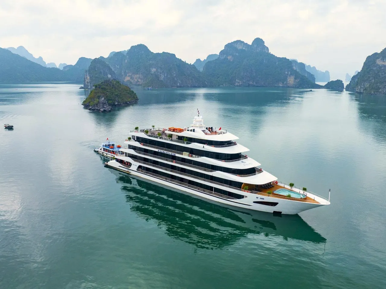 An aerial view of a large white multi-deck cruise ship with a small pool at the stern, sitting on calm green water among limestone karst islands, with a small boat in the distance and small lettering on the hull.