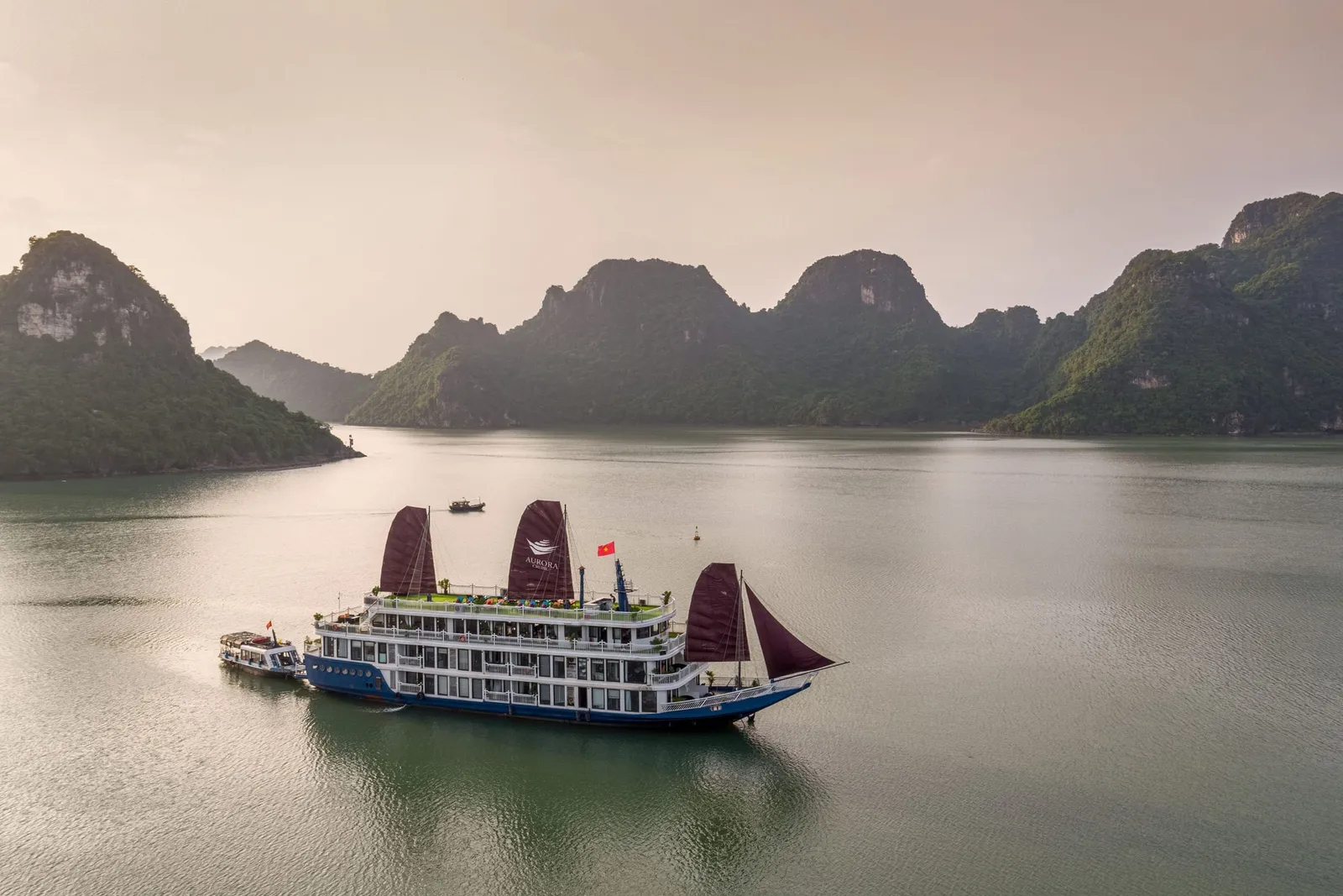 An aerial view of a multi-deck white and blue cruise boat with maroon sails, one bearing a logo, anchored on calm green water with a small tender boat beside it and forested limestone karst hills behind.