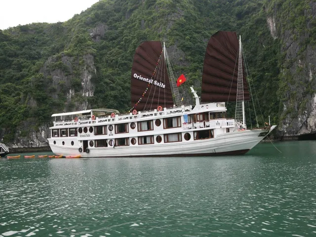 A white cruise junk with dark brown sails reading 'Oriental Sails' and a Vietnamese flag sits on green water in front of steep tree-covered limestone cliffs, with kayaks lined up beside it and another boat at left.