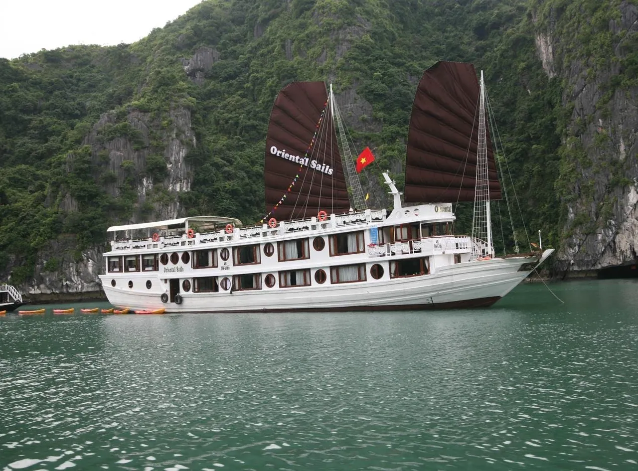 A white cruise junk with dark brown sails reading 'Oriental Sails' and a Vietnamese flag sits on green water in front of steep tree-covered limestone cliffs, with kayaks lined up beside it and another boat at left.