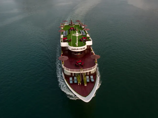 Aerial view of a multi-deck cruise boat with 'AMOUR CRUISE' lettered on its upper deck facade, moving through dark green water with a white wake, with umbrellas, lounge seating and sun loungers on its decks.