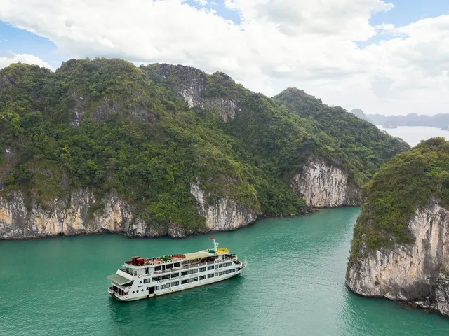 Aerial view of a white multi-deck cruise ship with 'La Regina Cruises' lettering on its hull sitting on emerald water between forested limestone karst cliffs under a cloudy blue sky.