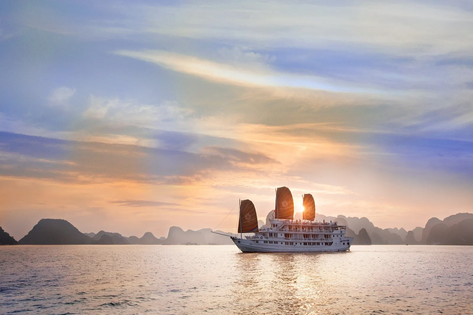 A white multi-deck cruise boat with three dark orange sails, some with a small emblem, floats on calm water at sunset with limestone karst silhouettes behind it.