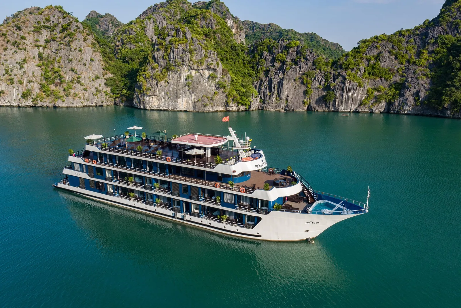 Aerial view of a white and blue multi-deck cruise ship with the name 'Rosy' on its hull and registration 'HP-5424' on the bow, sitting on green water in front of tall limestone karst cliffs covered in vegetation.
