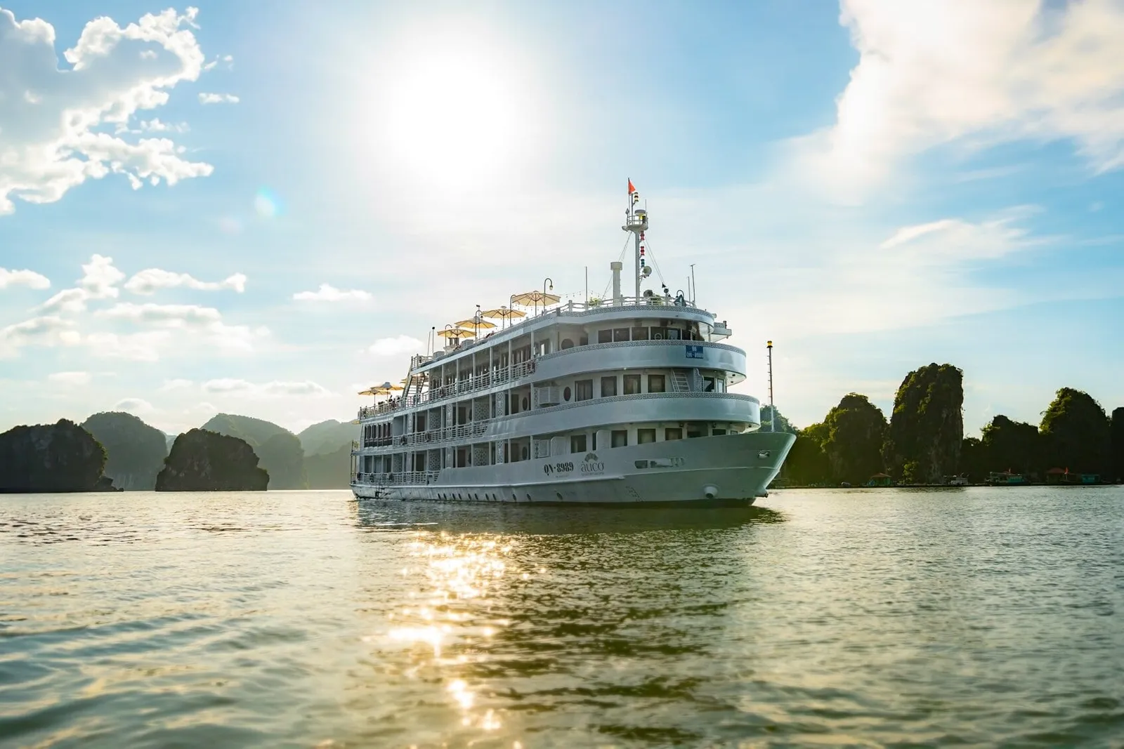 A large white multi-deck cruise ship with a flag and deck umbrellas sits on calm water in front of limestone karst islands under a bright sun and blue sky, with a logo and registration text painted on the hull.