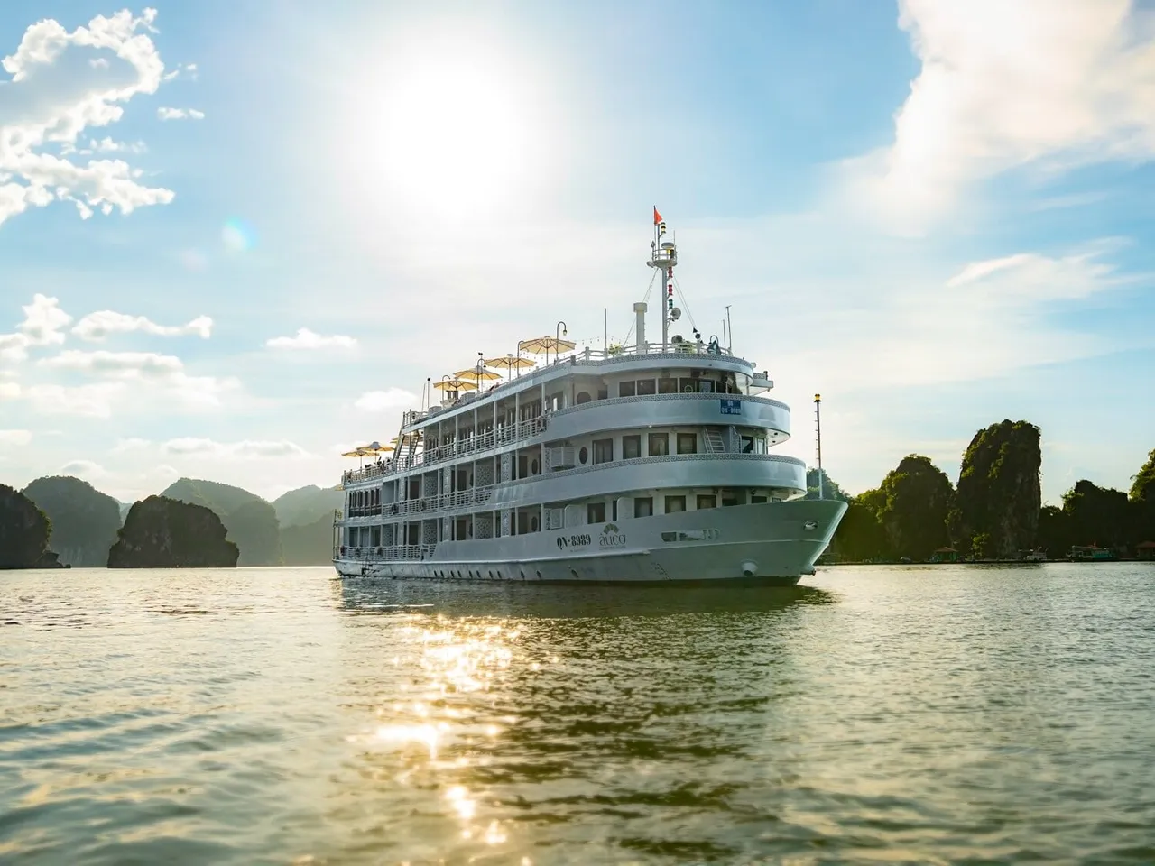 A large white multi-deck cruise ship with a flag and deck umbrellas sits on calm water in front of limestone karst islands under a bright sun and blue sky, with a logo and registration text painted on the hull.