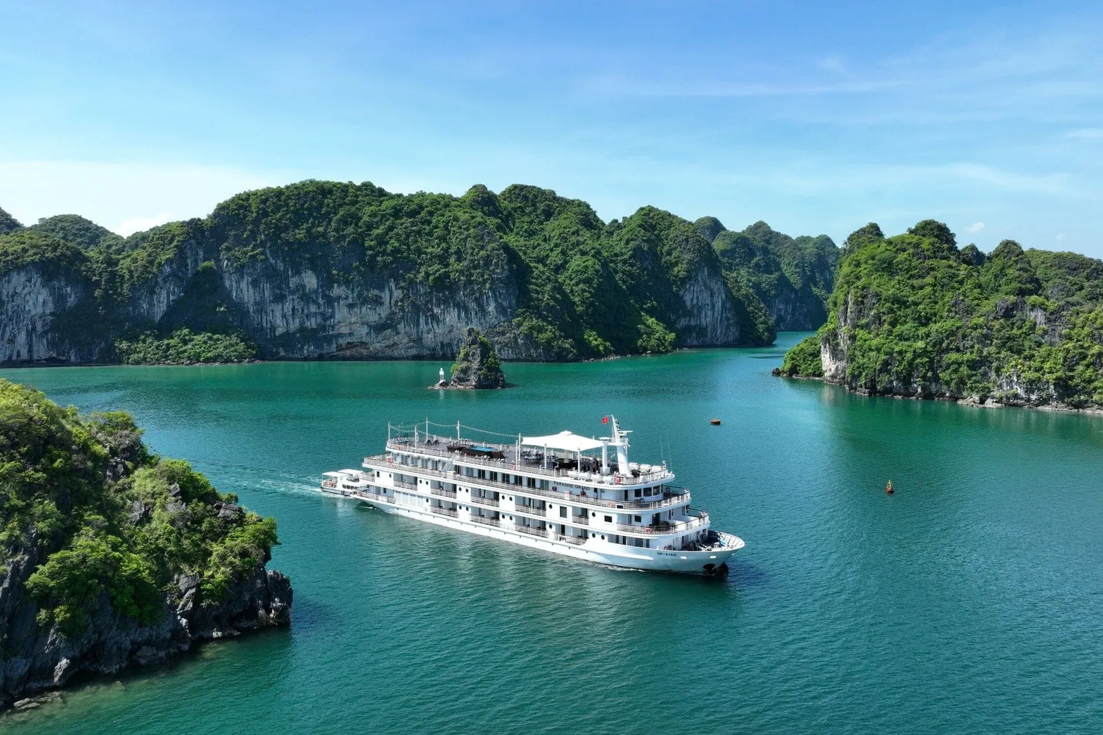 An aerial view of a multi-deck white cruise ship sailing on turquoise water between steep green limestone karst islands under a blue sky.