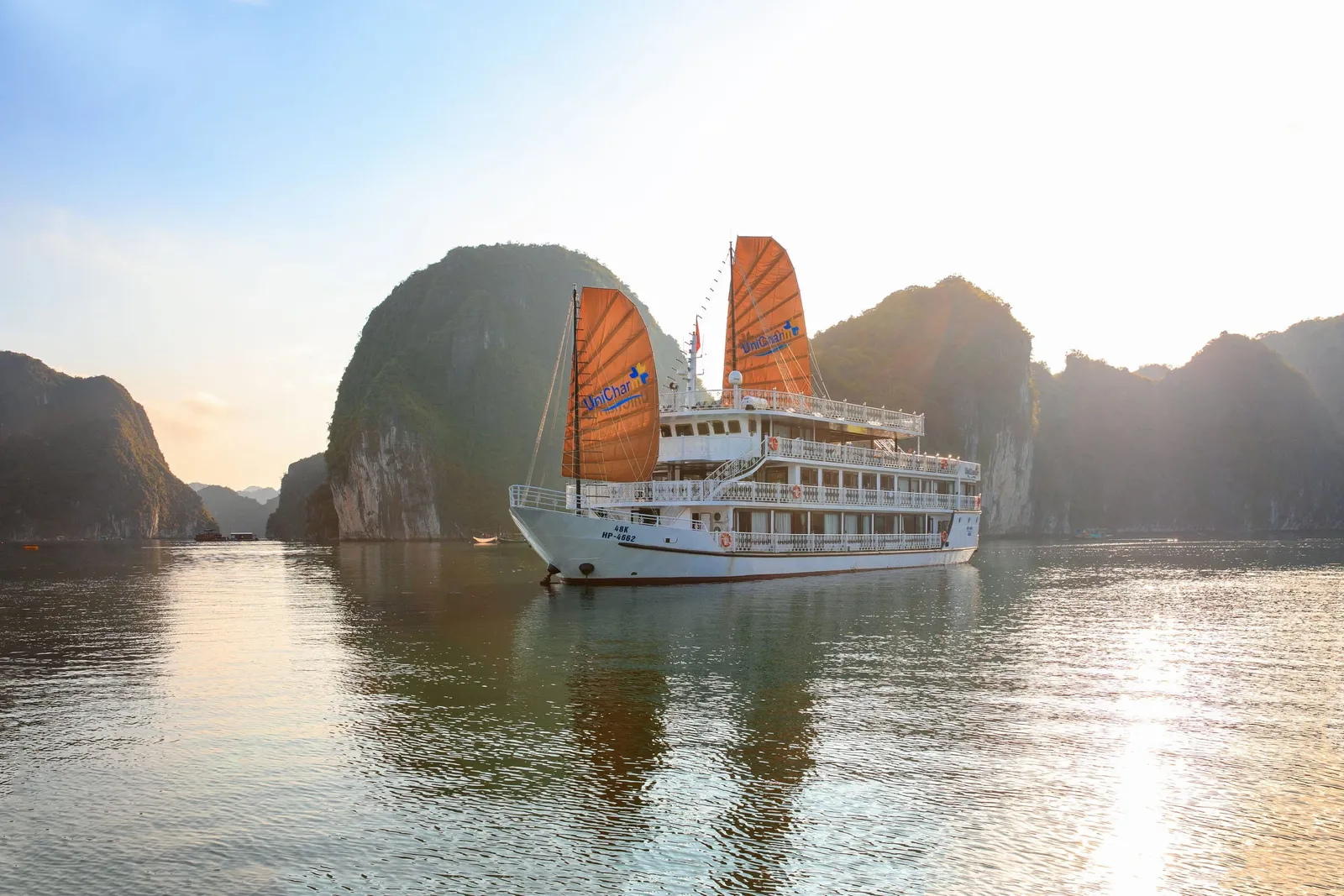 A white multi-deck cruise ship with orange sails bearing the name UniChar sits on calm water in front of tall limestone karst islands at sunset.