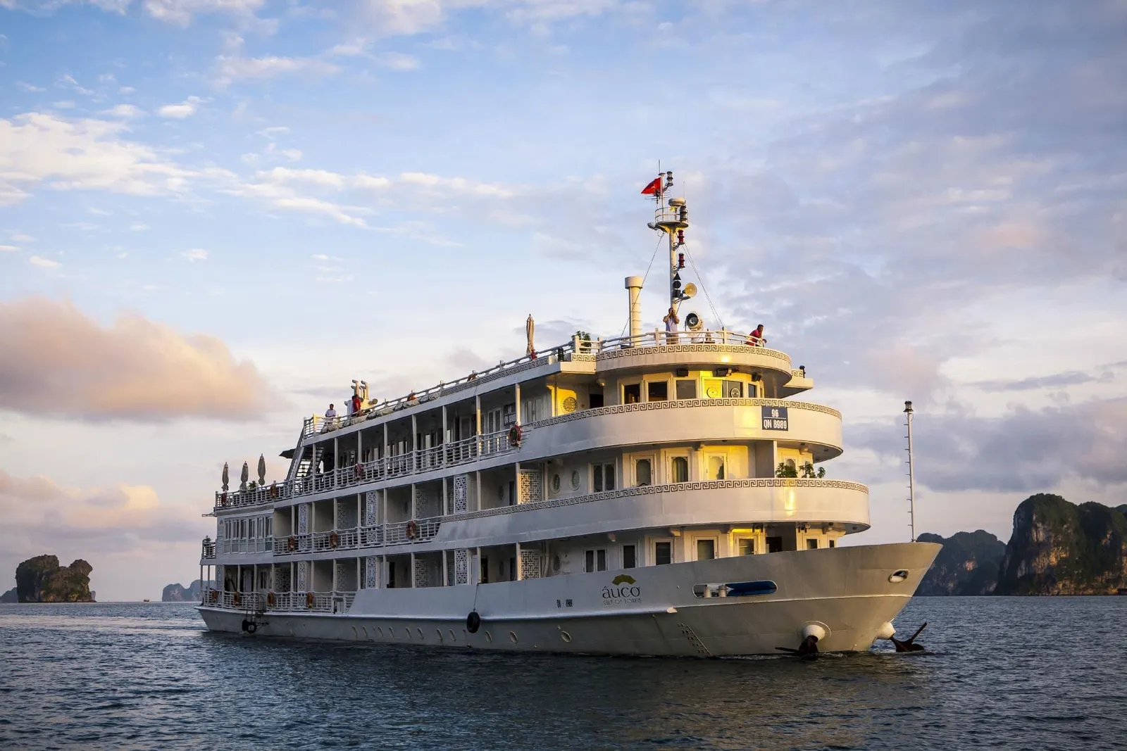 A multi-deck white cruise ship with the 'Auco' logo painted on its bow sails on calm water at sunset, with limestone karst islands in the background and a few people on the upper decks.