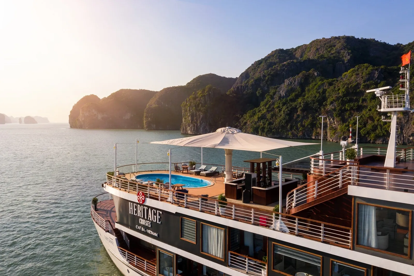 An elevated view of a cruise ship with a rooftop pool, sun deck and shade umbrella, with 'Heritage Cruises Cat Ba - Vietnam' painted on its hull, floating near forested limestone karsts in warm hazy light.