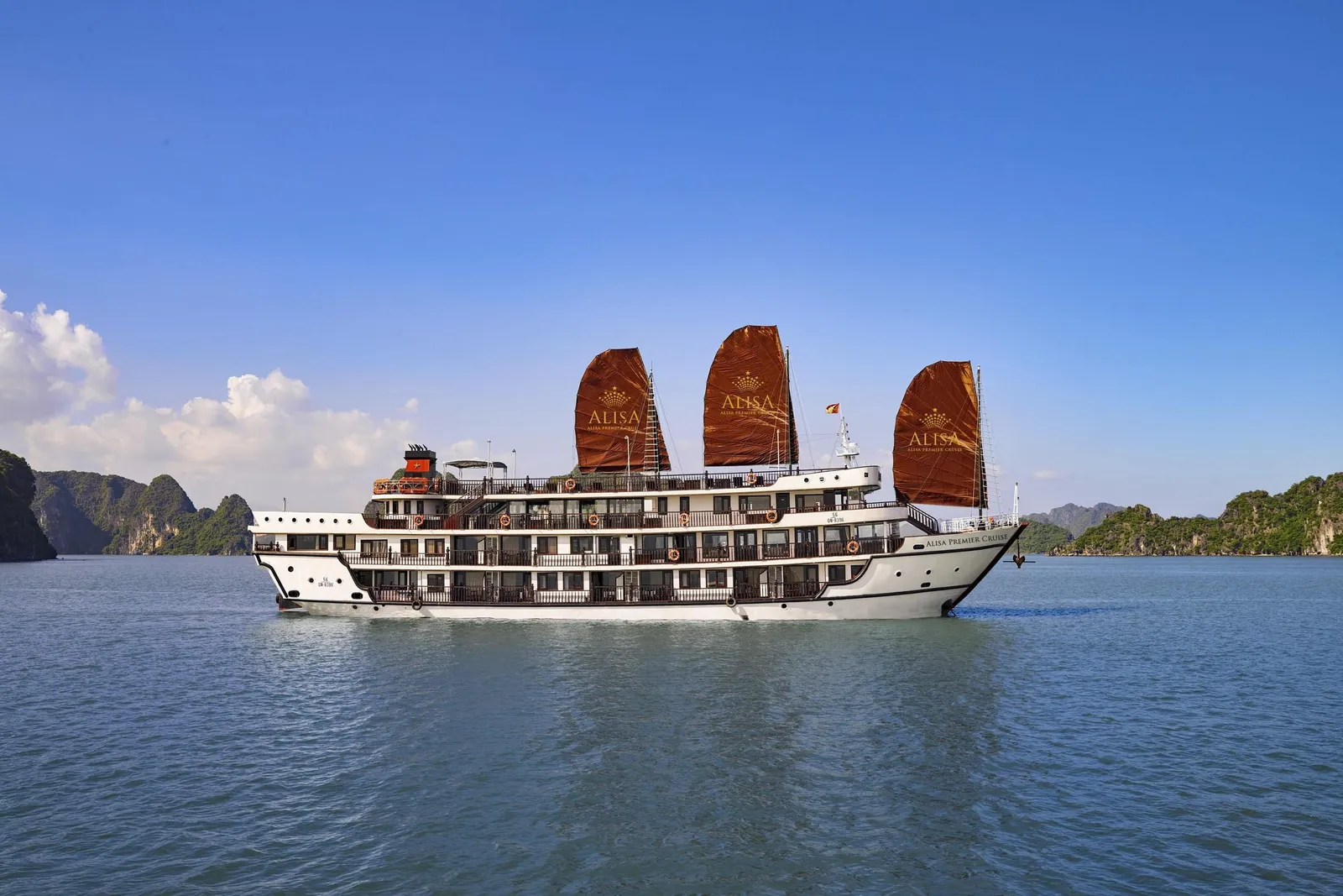 A white multi-deck cruise ship with three brown sails printed with the 'ALISA' name and crown logo, and 'Alisa Premier Cruise' on the hull, sails on calm blue water with limestone karst islands and a blue sky behind.