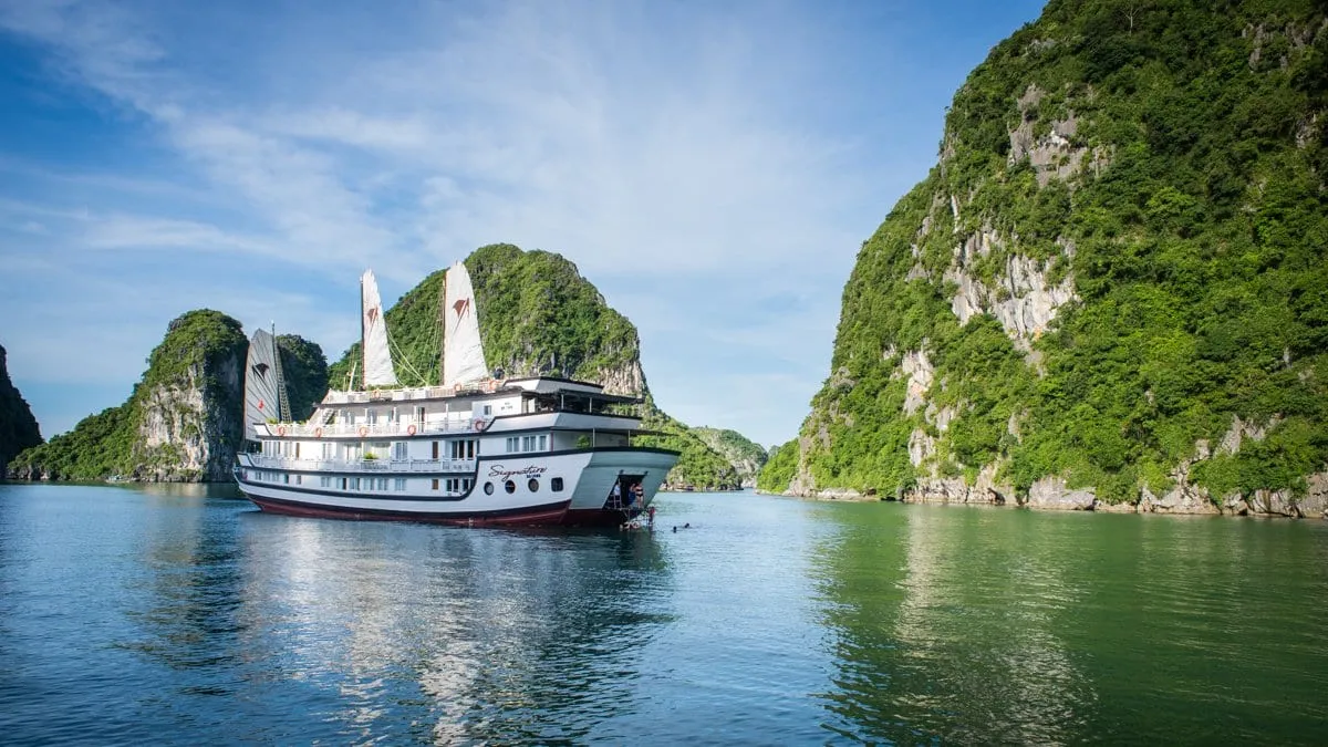A white multi-deck cruise boat with sails and the name "Signature" in script on its stern sits on calm green water between steep forested limestone karst islands under a blue sky.
