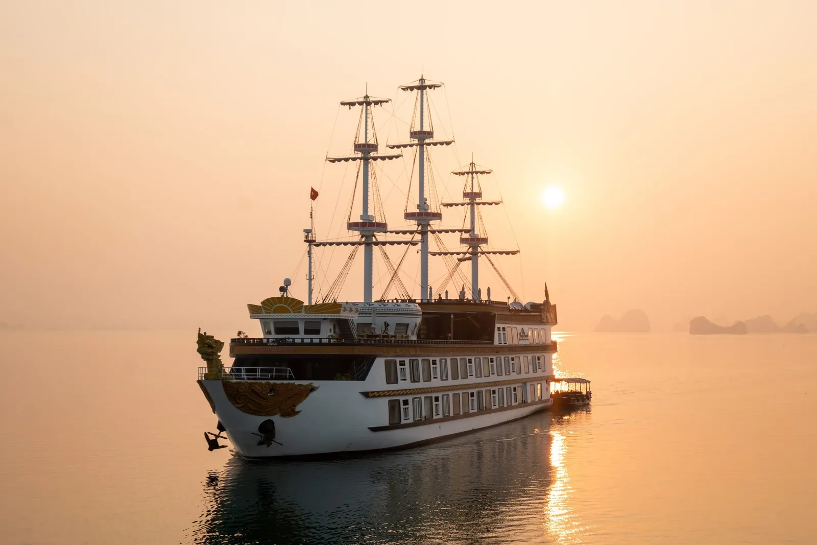 A white three-masted tall-ship-style cruise vessel with a gilded dragon bow ornament sits on calm water at sunset, with hazy limestone karst silhouettes in the distance.